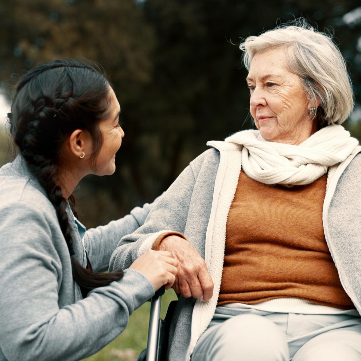Older Woman in Wheelchair Looks Down at Younger Adult Kneeling At Feet